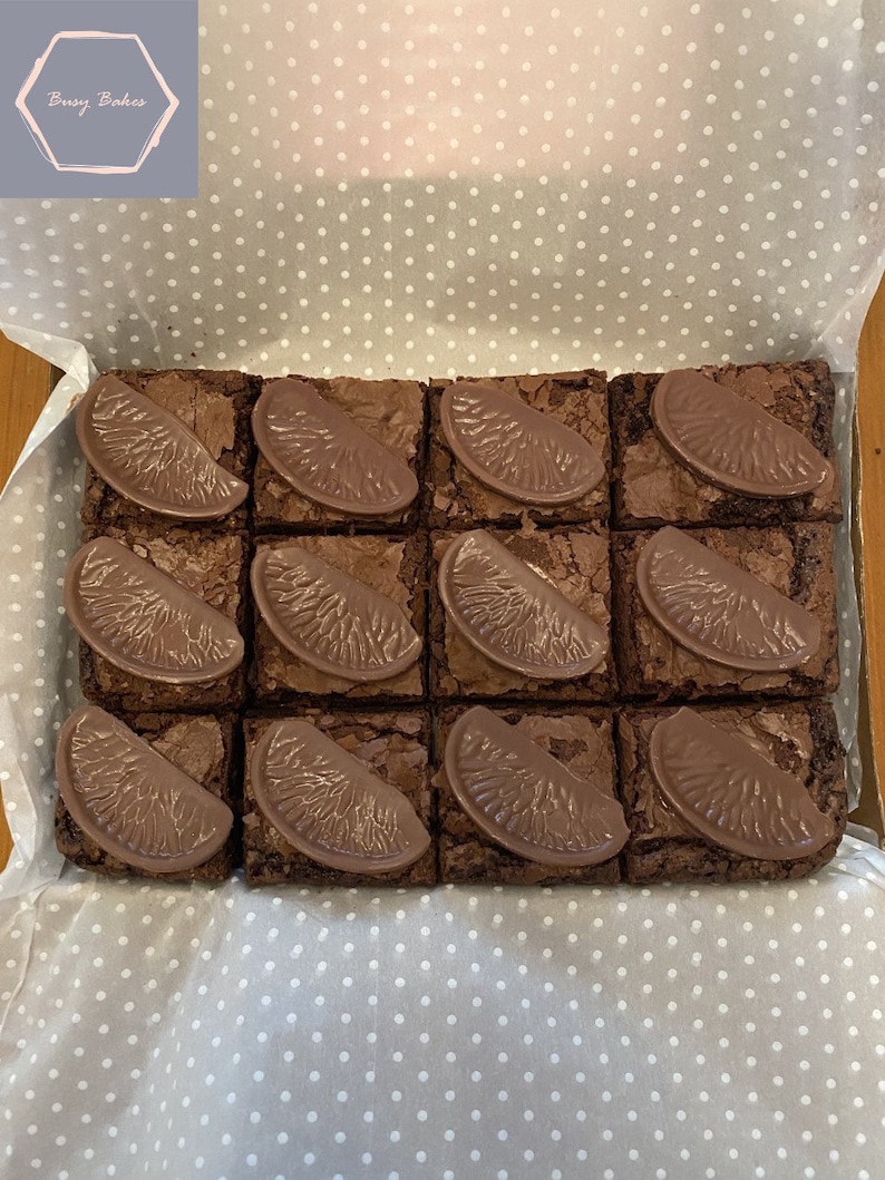 May include: A box of 16 chocolate brownies with orange slices on top. The brownies are arranged in a 4x4 grid. The box is lined with white paper with a polka dot pattern.