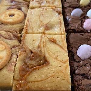 May include: A box of assorted brownies. The brownies are arranged in rows and include a variety of flavors, including caramel and chocolate. Some of the brownies are topped with candy.