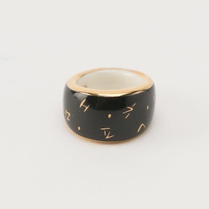 May include: A black ceramic ring with gold accents and a pattern of abstract symbols.