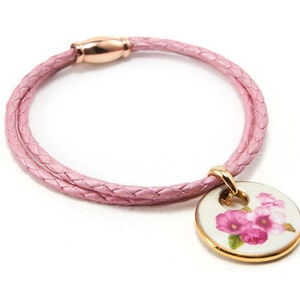 May include: A pink leather bracelet with a braided design and a gold charm featuring a floral pattern.