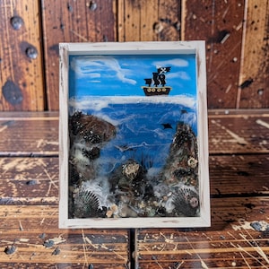 May include: A framed nautical scene featuring a pirate ship sailing on a blue ocean under a blue sky with white clouds. The artwork includes rocks, seashells, and a white frame. The pirate ship has a black flag with a skull and crossbones.
