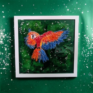 May include: A colorful parrot painting in a white frame. The artwork features a vibrant parrot with red, orange, and blue feathers against a green background. The painting is square and appears to be made with textured paint.