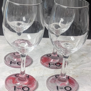 May include: Four clear glass wine glasses with stems and bases featuring a marbled pink and white resin design. Each base is marked with the black text "I-O" and "H-O" printed on the surface, arranged in a set of four identical drinking vessels.