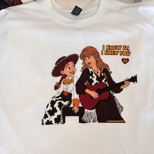 May include: A white t-shirt featuring a graphic print of a cowgirl character sitting next to a woman with long wavy hair playing an acoustic guitar. The text reads I KNEW IT, I KNEW YOU with a heart icon containing the letters TS above the guitar.