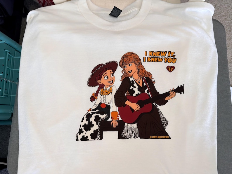 May include: A white t-shirt featuring a graphic print of a cowgirl character sitting next to a woman with long wavy hair playing an acoustic guitar. The text reads I KNEW IT, I KNEW YOU with a heart icon containing the letters TS above the guitar.
