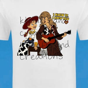May include: A white t-shirt featuring a graphic illustration of a cowgirl character with red hair and a blonde woman playing an acoustic guitar. The text "I KNEW IT, I KNEW YOU" is printed in yellow and black, with a small red heart containing the letters TS.