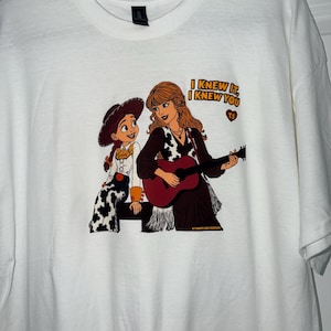May include: A white t-shirt featuring a graphic print of a cartoon cowgirl and a woman with long hair playing a guitar. The text "I KNEW IT, I KNEW YOU" is printed in yellow above a small red heart containing the letters "TS" and "KT CRAFTS AND CREATIONS".