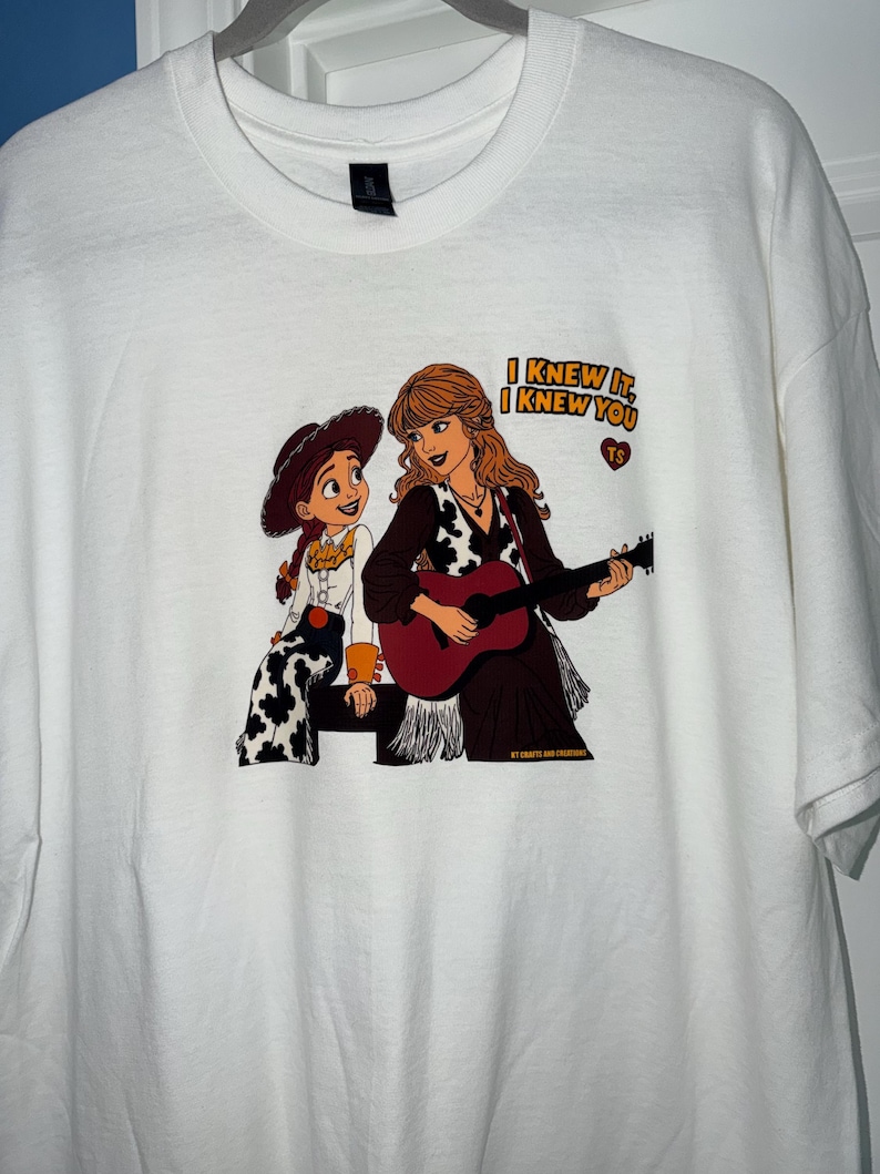 May include: A white t-shirt featuring a graphic print of a cartoon cowgirl and a woman with long hair playing a guitar. The text "I KNEW IT, I KNEW YOU" is printed in yellow above a small red heart containing the letters "TS" and "KT CRAFTS AND CREATIONS".