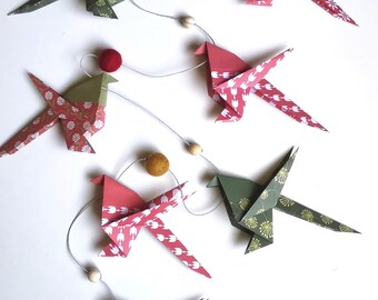Może przedstawiać: Girlanda z ptaków origami w odcieniach czerwieni, zieleni i bieli, z kwiatowymi wzorami. Każdy ptak połączony jest cienkim sznurkiem, przeplatanym małymi drewnianymi koralikami i filcowymi kulkami. Ptaki ułożone są na białym tle.