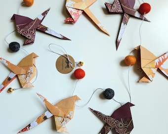 Może przedstawiać: Girlanda z ptaków origami w odcieniach pomarańczu i bordo, z akcentami z filcowych kulek. Ptaki są połączone cienką nitką, a niektóre mają małe dzwoneczki. Widoczna jest ręcznie robiona etykieta.