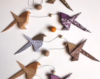 Może przedstawiać: Girlanda z ptaków origami w różnych kolorach i wzorach, w tym brązowym, fioletowym i złotym, połączonych sznurkiem, drewnianymi koralikami i filcowymi kulkami. Ptaki mają kwiatowe i geometryczne wzory. Dołączona jest mała etykieta z napisem "handmade".