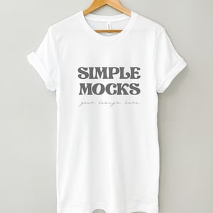 Può includere: Maglietta bianca con il testo "SIMPLE MOCKS" in un font retrò e le parole "your design here" scritte in corsivo sotto.