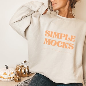 Puede incluir: Una sudadera blanca con el texto "SIMPLE MOCKS" en letras naranjas. La sudadera la lleva puesta una persona con el pelo largo y castaño. En primer plano hay tres calabazas, una blanca, una dorada y una pequeña calabaza naranja.