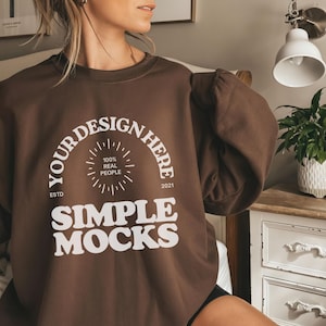 Puede incluir: Una sudadera marrón con el texto "YOUR DESIGN HERE" en un diseño circular, con "SIMPLE MOCKS" debajo. La sudadera tiene mangas largas y cuello redondo. El texto también incluye "100% REAL PEOPLE" y el año "2021".
