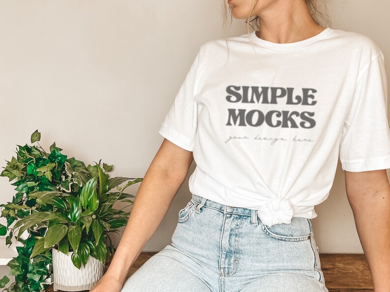 May include: A white t-shirt with the text "SIMPLE MOCKS" in a retro font. The text is in gray and the shirt is tied at the waist. The person wearing the shirt is wearing blue jeans.