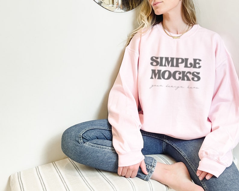 May include: A pink sweatshirt with the text "SIMPLE MOCKS your design here" in black. The sweatshirt is being worn by a person sitting on a striped surface.