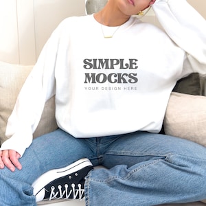 Może przedstawiać: Biała bluza z napisem "SIMPLE MOCKS YOUR DESIGN HERE" w kolorze czarnym. Bluzę nosi osoba siedząca na kanapie w niebieskich dżinsach i czarnych butach sportowych.