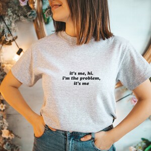 May include: A gray t-shirt with the text "it's me, hi. i'm the problem, it's me" printed on the front.