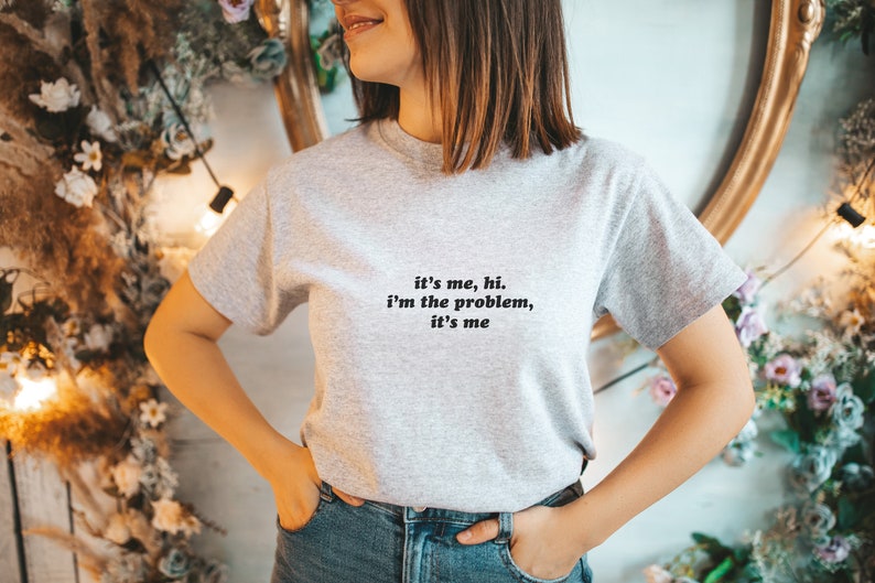 May include: A gray t-shirt with the text "it's me, hi. i'm the problem, it's me" printed on the front.