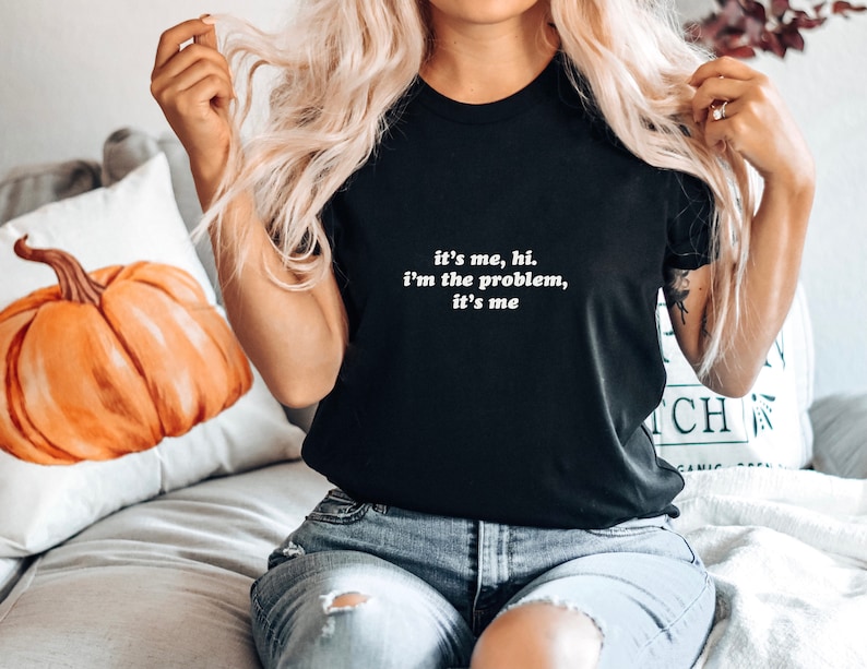 May include: A black t-shirt with white text that reads "it's me, hi. i'm the problem, it's me".