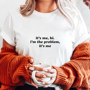 May include: White t-shirt with the text "it's me, hi. i'm the problem, it's me" printed on the front. The person is wearing a brown sweater and holding a white mug.