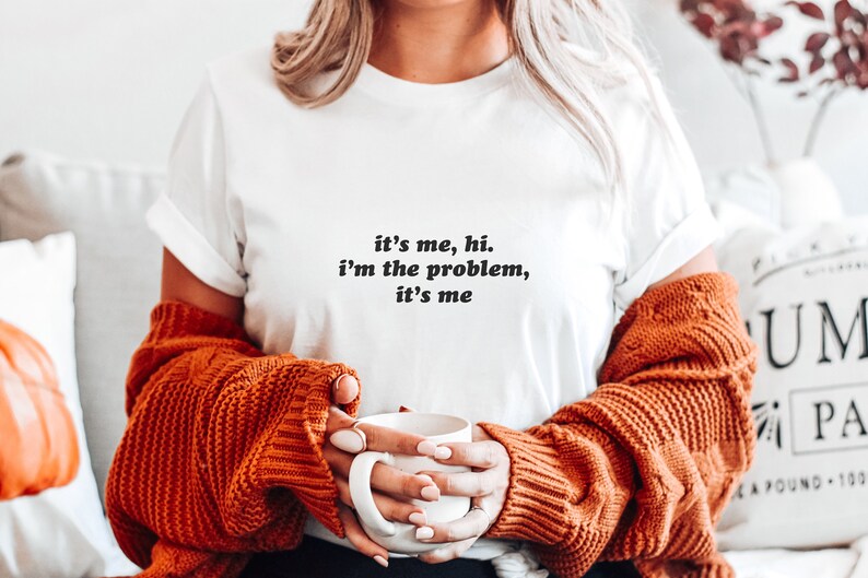 May include: White t-shirt with the text "it's me, hi. i'm the problem, it's me" printed on the front. The person is wearing a brown sweater and holding a white mug.