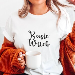 May include: A white t-shirt with the words "Basic Witch" in black script. The person is wearing a burnt orange sweater and holding a white mug. The image has a cosy, autumnal theme.