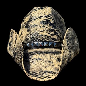 May include: A cowboy hat with a snakeskin pattern in beige and black. The hat features a black band adorned with silver pyramid-shaped studs. The hat's brim is curved, and the background is black.