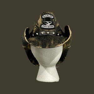 May include: A camouflage cowboy hat with a black leather band and silver studs. The hat features a Harley-Davidson patch with a skull and the brand name in white lettering. The hat is displayed on a white mannequin head.