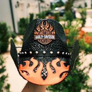 Puede incluir: Un sombrero de vaquero negro trenzado con un diseño de llamas naranjas en el ala delantera y la copa. Una banda de cuero negro con tachuelas metálicas plateadas rodea la base de la copa. El logotipo de Harley-Davidson Motor Cycles está centrado.