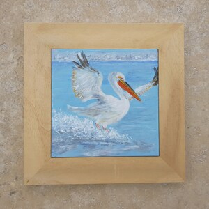 May include: A framed painting depicting a white pelican with an orange beak taking off from the water. The pelican is in mid-flight with its wings spread wide. The water is a light blue colour and there are white splashes of water around the pelican.