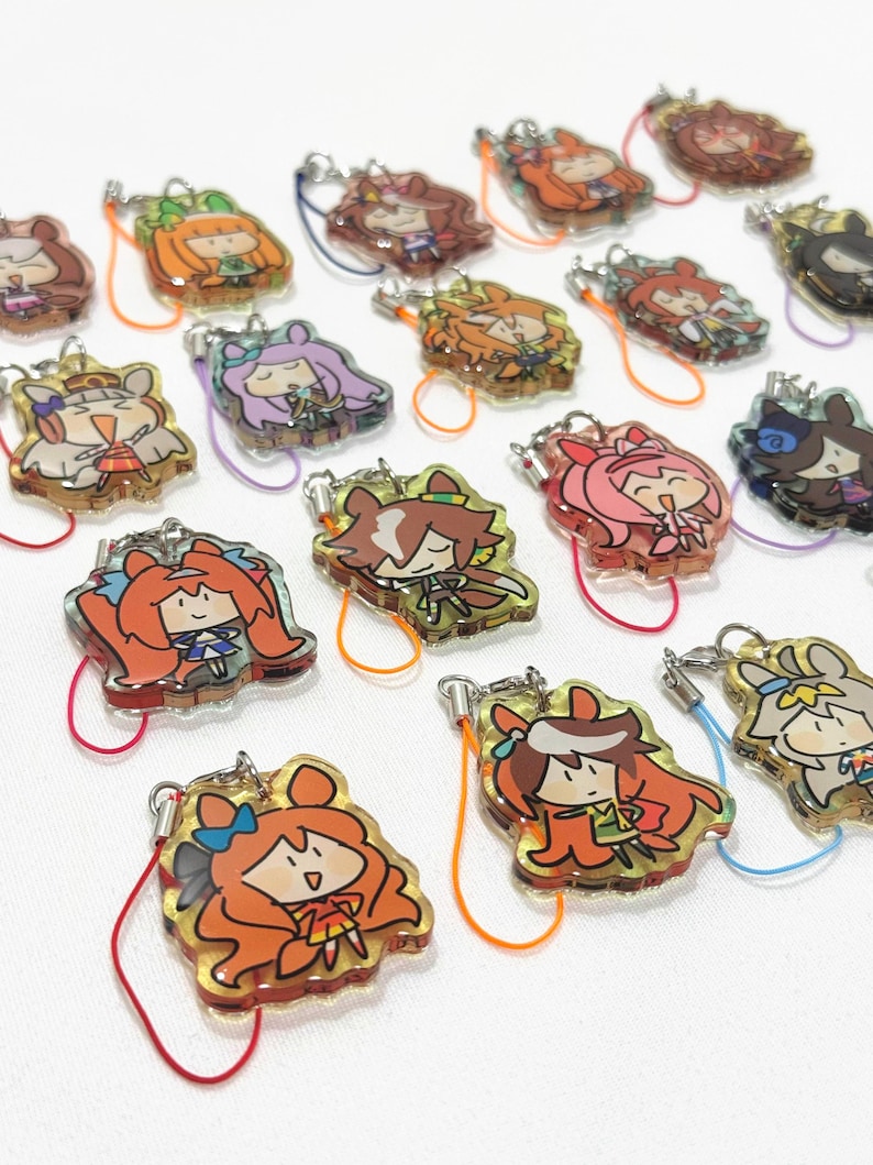 以下が含まれることがあります： アニメスタイルのキャラクターデザインが施された、カラフルなアクリルチャームのコレクション。各チャームには、さまざまな髪の色と衣装を持つユニークなキャラクターがおり、カラフルなストラップが付いたシルバーカラーの留め金が付いています。