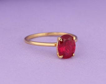 May include: A gold ring with a large, oval-shaped, red gemstone. The ring is set against a soft purple background, creating a striking contrast. The gemstone is the focal point, showcasing its vibrant color and brilliance.