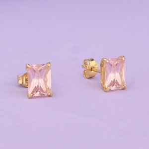 May include: A pair of gold-colored stud earrings, each featuring a rectangular, light pink gemstone. The earrings are set against a soft purple background, showcasing their elegant design and color.