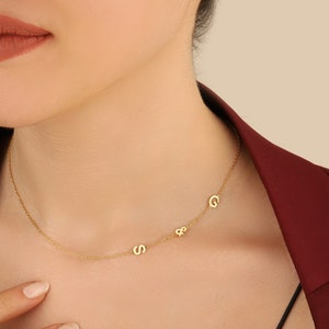 May include: A gold chain necklace with three small letters, "S", "&", and "C", hanging from it. The necklace is worn by a person with a burgundy blazer.