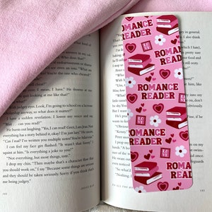 Peut inclure: Signet de livre rose et rouge avec une pile de livres et le texte "Romance Reader" répété partout. Le signet a un fond rose avec des fleurs blanches et des cœurs rouges.
