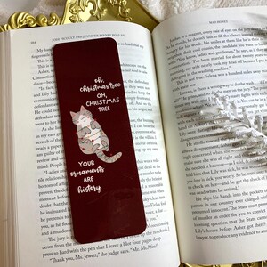 May include: A red bookmark featuring a white cat adorned with Christmas lights. The bookmark includes the text "Oh, Christmas tree, Oh, Christmas tree. Your ornaments are history."