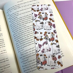 May include: A white bookmark featuring a repeating pattern of ghosts and flowers. The ghosts are white with black outlines and are holding flowers. The flowers are in shades of pink, yellow, and orange.