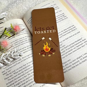 May include: A brown bookmark featuring the text "lets get TOASTED" and a campfire illustration. The design includes a campfire with flames, logs, and marshmallows on sticks. The bookmark is on an open book.