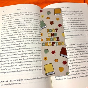 Peut inclure: Marque-page rectangulaire avec l'inscription "JUST ONE MORE CHAPTER" et des illustrations d'automne. Le marque-page est inséré dans un livre ouvert. On y voit des livres, des tasses à café, des citrouilles et des feuilles d'érable.