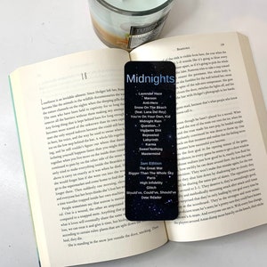 May include: A black and white bookmark with the title "Midnights" and a list of song titles from the album. The bookmark is open to the page with the song titles "Lavender Haze", "Maroon", "Anti-Hero", "Snow On The Beach", "You're On Your Own, Kid", "Midnight Rain", "Question?", "Vigilante Shit", "Bejeweled", "Labyrinth", "Karma", "Sweet Nothing", "Mastermind", "3am Edition", "The Great War", "Bigger Than The Whole Sky", "High Infidelity", "Glitch", "Would've, Could've, Should've", "Dear Reader".
