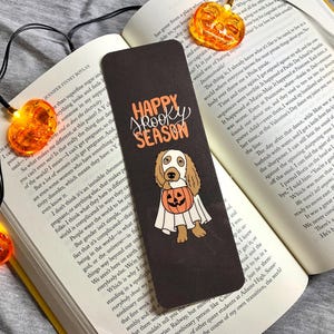 May include: A black bookmark featuring a white ghost dog holding a jack-o'-lantern. The bookmark has the text "Happy Spooky Season" in white lettering.