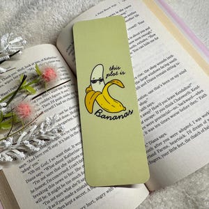 May include: A pale green bookmark featuring a cartoon banana in sunglasses with the phrase "this plot is bananas". The bookmark rests on an open book with text, alongside white floral decorations.