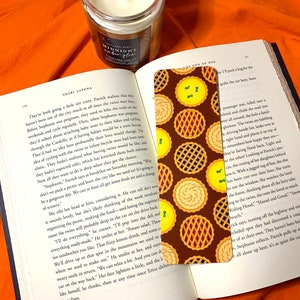 Peut inclure: Gros plan d'un marque-page brun avec un motif de tranches de tarte jaunes et brunes. Le marque-page est placé sur un livre ouvert avec le texte "The End of Me" visible sur la page.