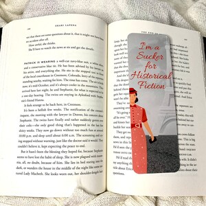 May include: A bookmark with a gray background and a red and white illustration of a woman in a red dress and a red hat standing in front of the Eiffel Tower. The text "I'm a Sucker for Historical Fiction" is written in black text on the bookmark.