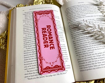 Puede incluir: Marcapáginas rosa con el texto "ROMANCE READER" en rojo, colocado en un libro abierto. El marcapáginas tiene un borde rojo decorativo con un diseño de encaje. Las páginas del libro son visibles.