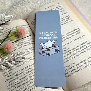 Puede incluir: Un marcapáginas azul claro con el texto "Crying Over Fictional Characters" y una ilustración de una caja de pañuelos. El marcapáginas está sobre un libro abierto con detalles florales.