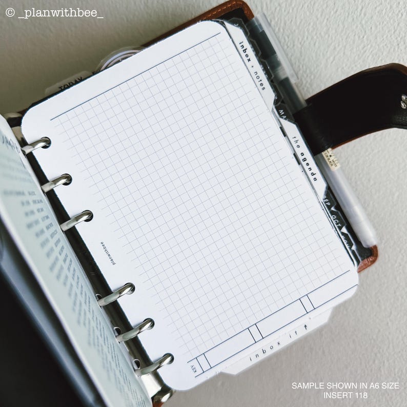 Op de afbeelding: Een plannerinzet met een rasterpatroon, getoond in een open binder. De inzet heeft gelabelde tabbladen voor 'inbox notes' en 'the agenda'. De tekst 'SAMPLE SHOWN IN A6 SIZE INSERT 118' is zichtbaar. De planner is bruin en zwart.