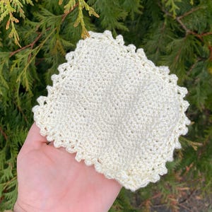 May include: A square, cream-coloured crochet coaster with a scalloped edge. The handmade coaster has a textured surface. The background is a blurred view of green foliage.