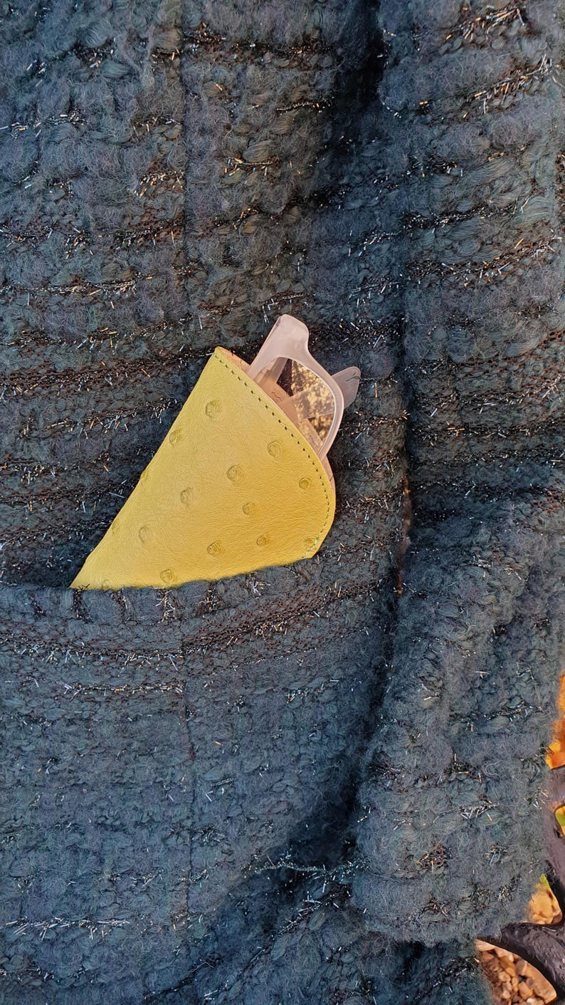 May include: A yellow, textured, semi-circular glasses case with visible stitching along the curved edge, containing a pair of light-colored eyeglasses, tucked into the pocket of a dark teal, chunky, metallic-flecked knit jacket.